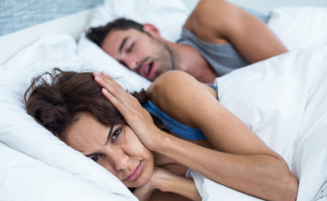 woman blocking ears with hands while man snoring on bed
